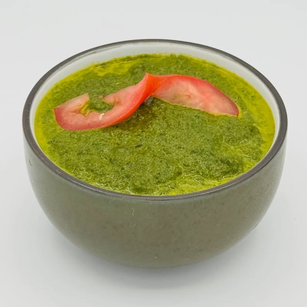 A bowl of vibrant green Indian chutney, garnished with two slices of red tomato.