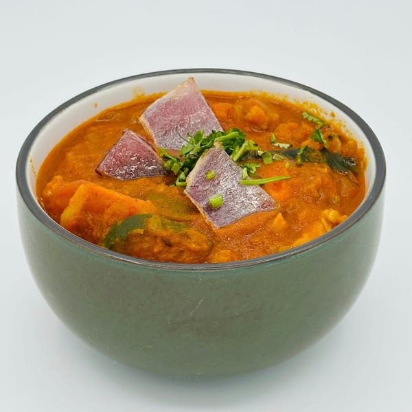 A vibrant orange Indian curry with purple yam and green herbs in a grey bowl.